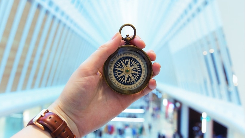 A hand holds up a brass compass against the backdrop of a modern glass building's soaring, symmetrical architecture.